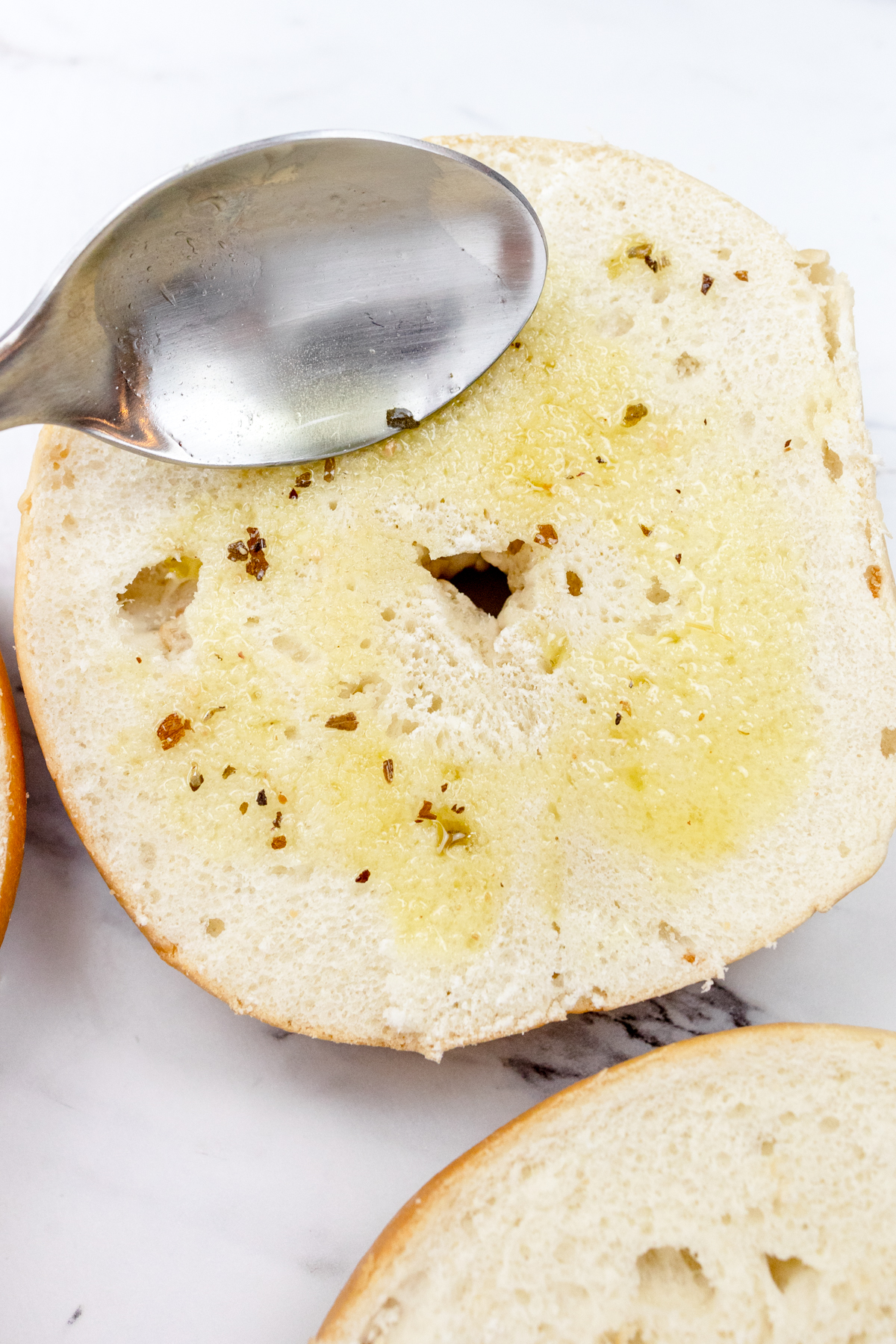 Close up view of bagel slices with oil and Italian Seasonings being spread on them by a spoon.