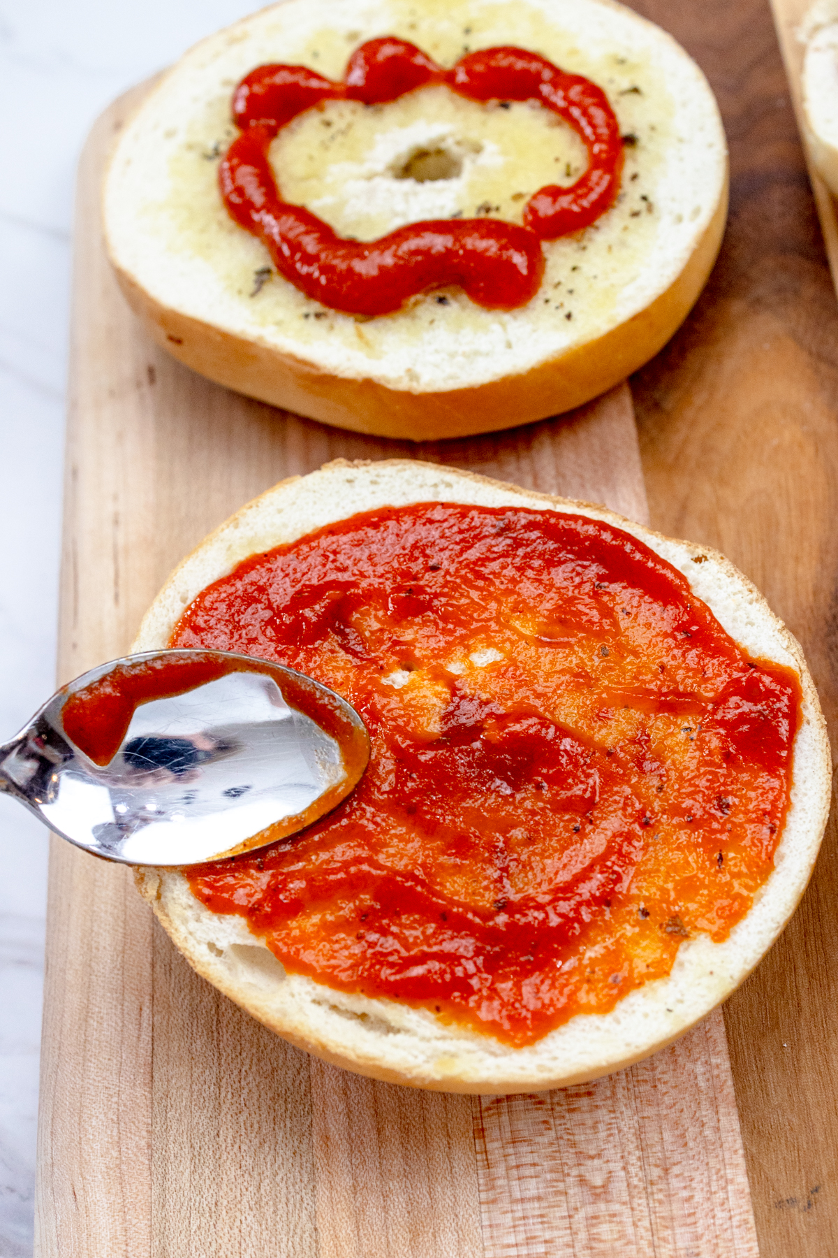 Close up view of bagel slices with pizza sauce being spread on them with a spoon.