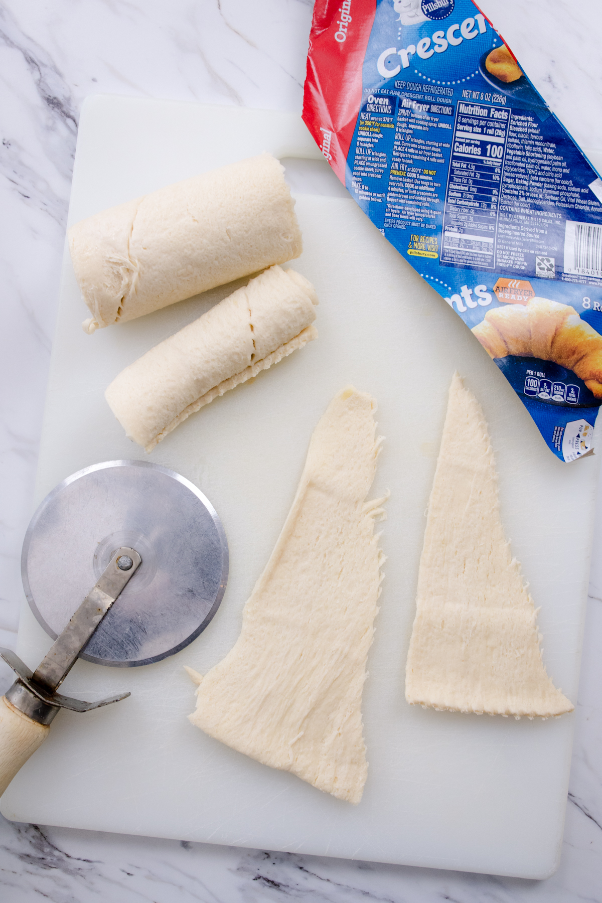 Top view of a white chopping board on a marble surface which has crescent rolls pastry next to a pizza cutter.