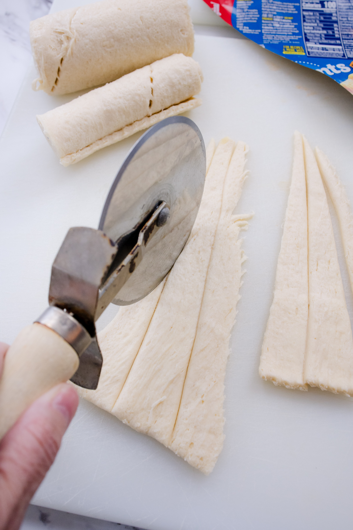 Close up view view of a white chopping board on a marble surface which has crescent rolls pastry being sliced by a pizza cutter.