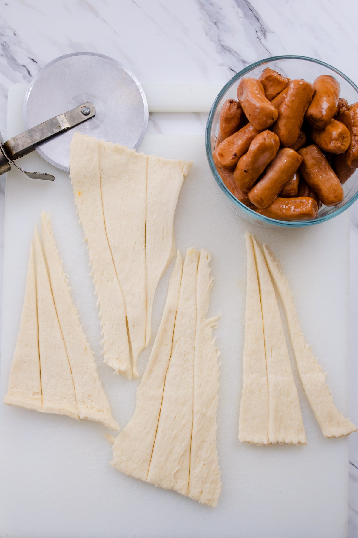 Top view of a white chopping board on a marble surface which has sliced crescent rolls pastry next to a pizza cutter and a bowl of Lil Smokies.