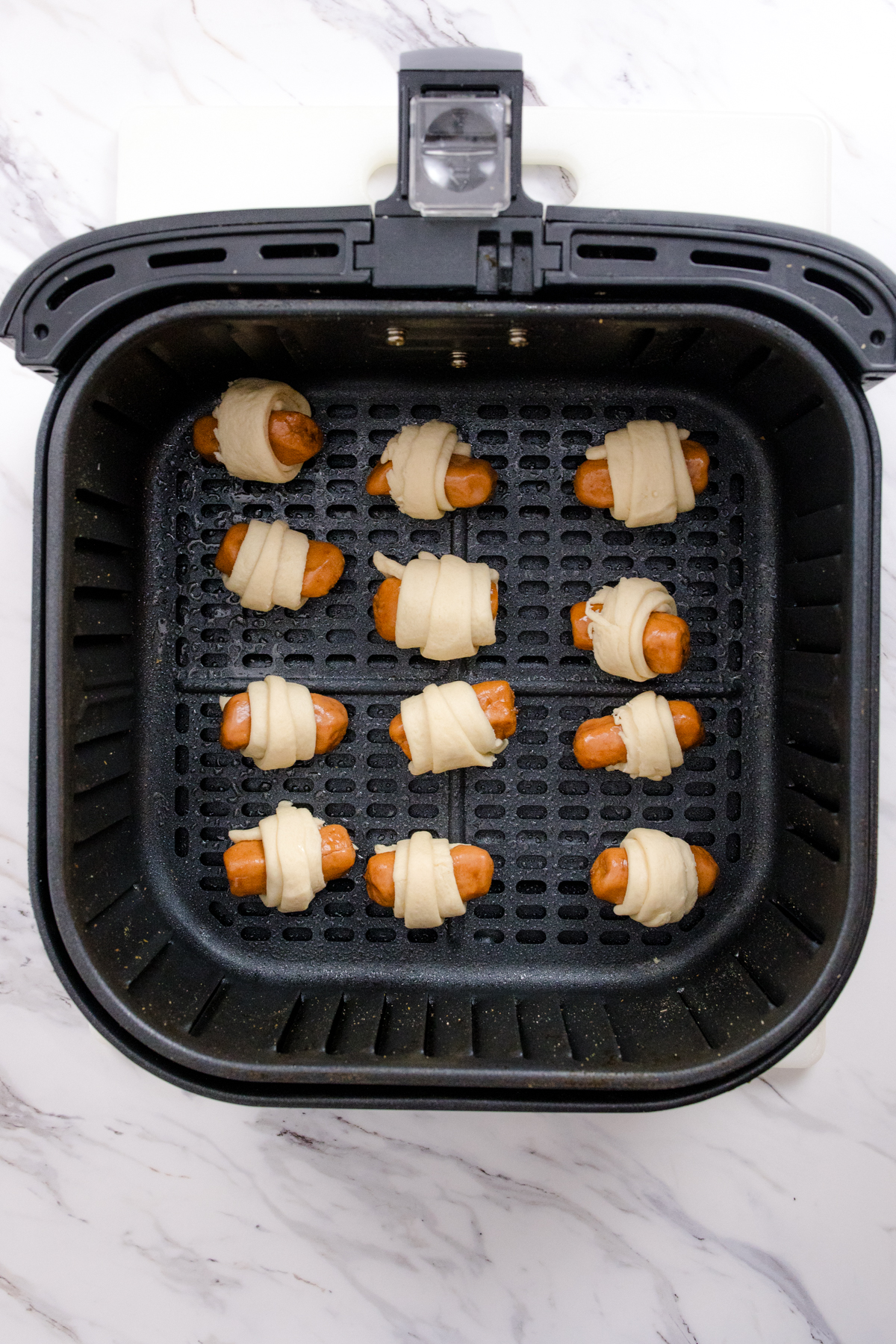 Top view of 12 uncooked Pigs in a Blanket in an air fryer basket.