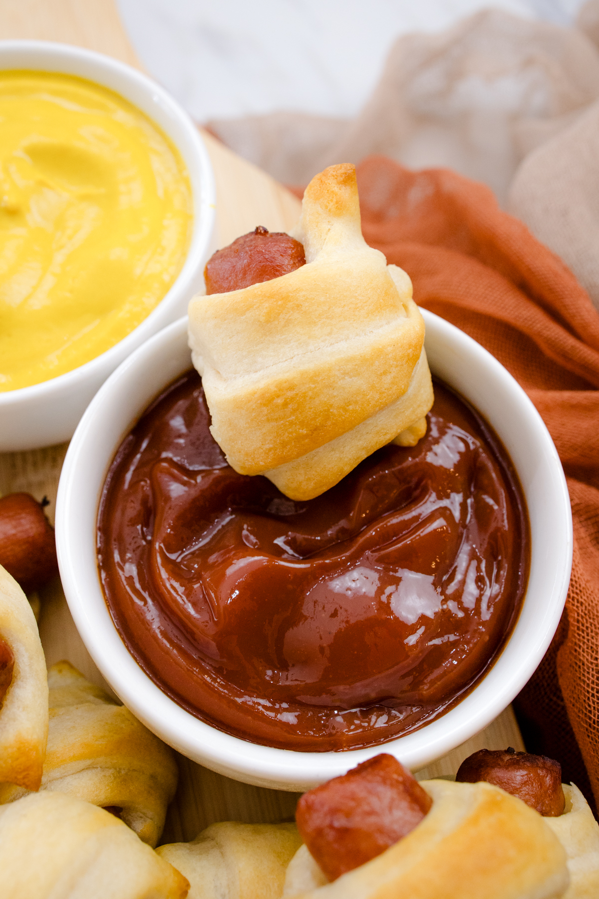 Close up view of an air fried Pig in a Blanket being dipped into a ramekin of ketchup, on a countertop with decorations and other pigs in blankets, next to a ramekin filled with mustard.