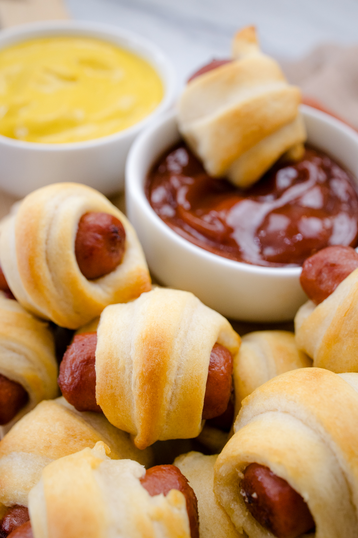 Close up view of air fried Pigs in a Blanket on a countertop with decorations next to ramekins filled with tomato ketchup and mustard. One pig in a blanket is in the ketchup ramekin.