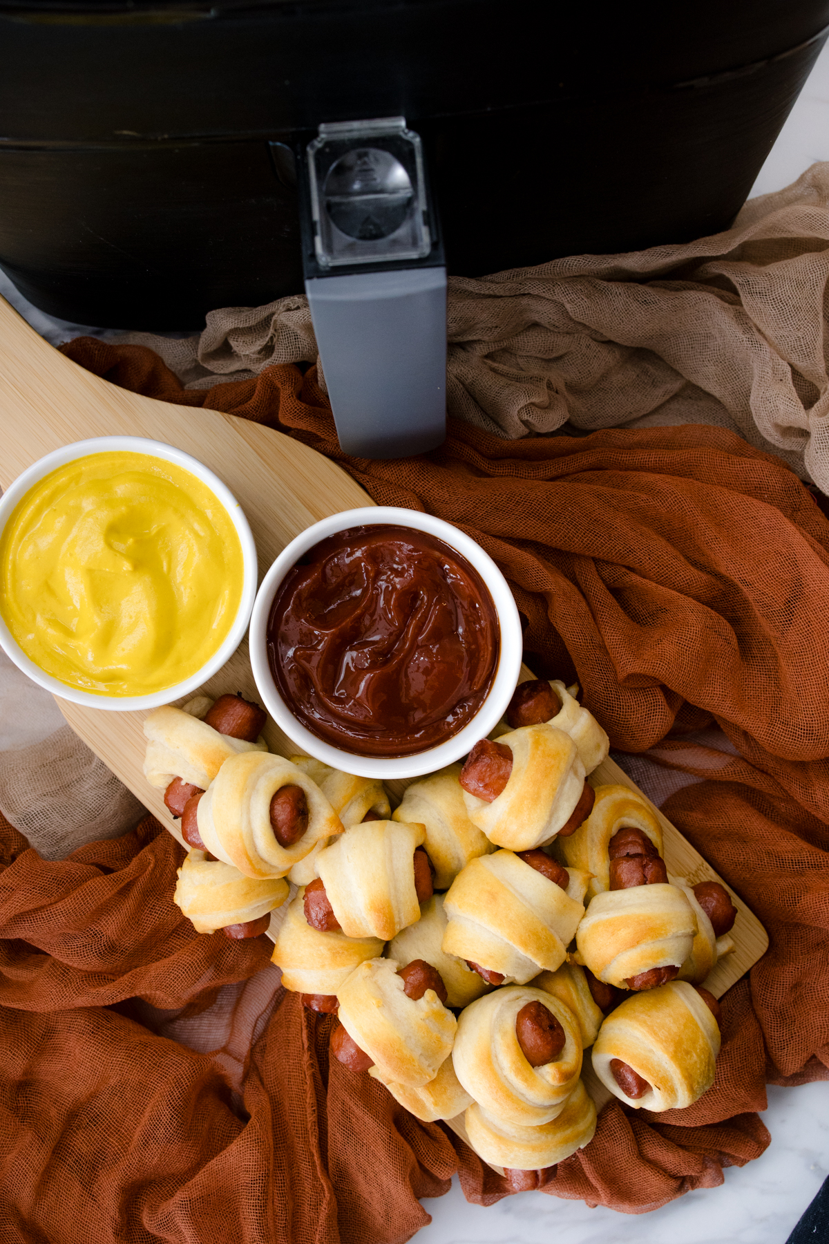 Top view of air fried Pigs in a Blanket on a countertop with decorations next to ramekins filled with tomato ketchup and mustard in front of an air fryer.