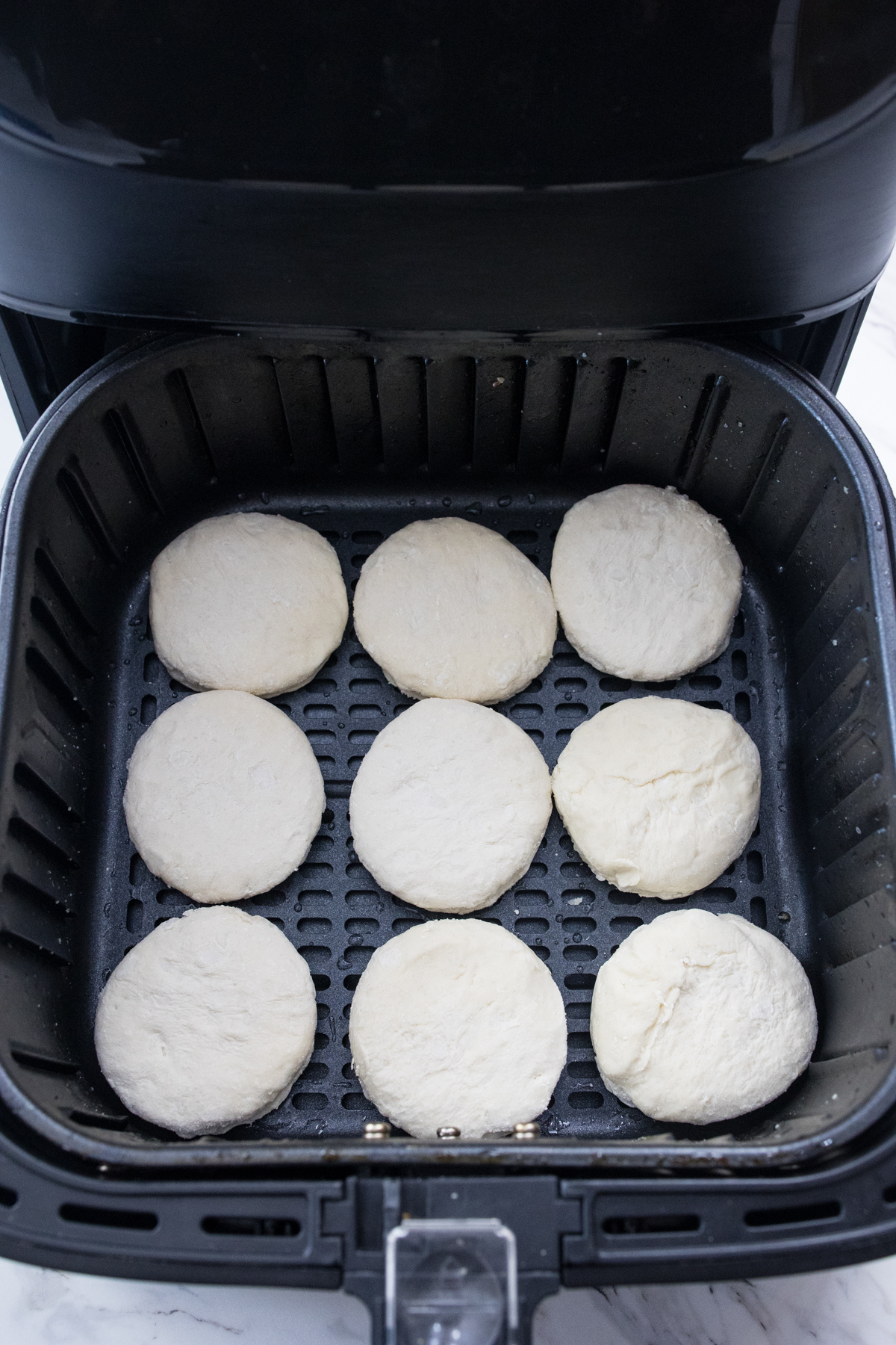 Top view of an open air fryer with frozen biscuits in it.