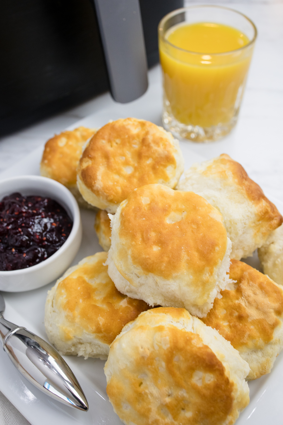 Close view of cooked biscuits on a white surface next to a ramakin of jam and a glass of orange juice.