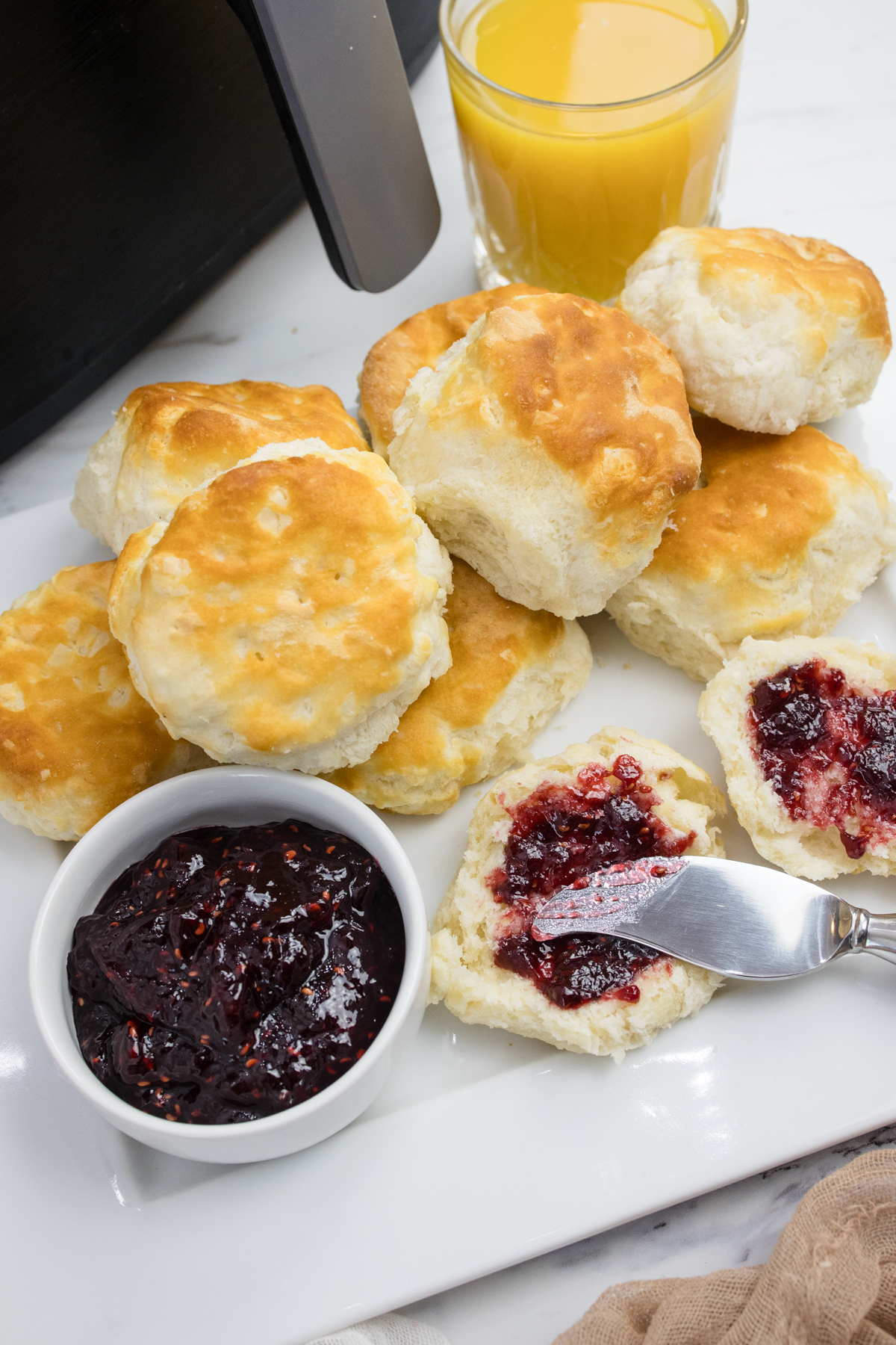 Close view of cooked biscuits on a white surface next to a ramakin of jam and a glass of orange juice. A knife is spreading jam onto a sliced biscuit.