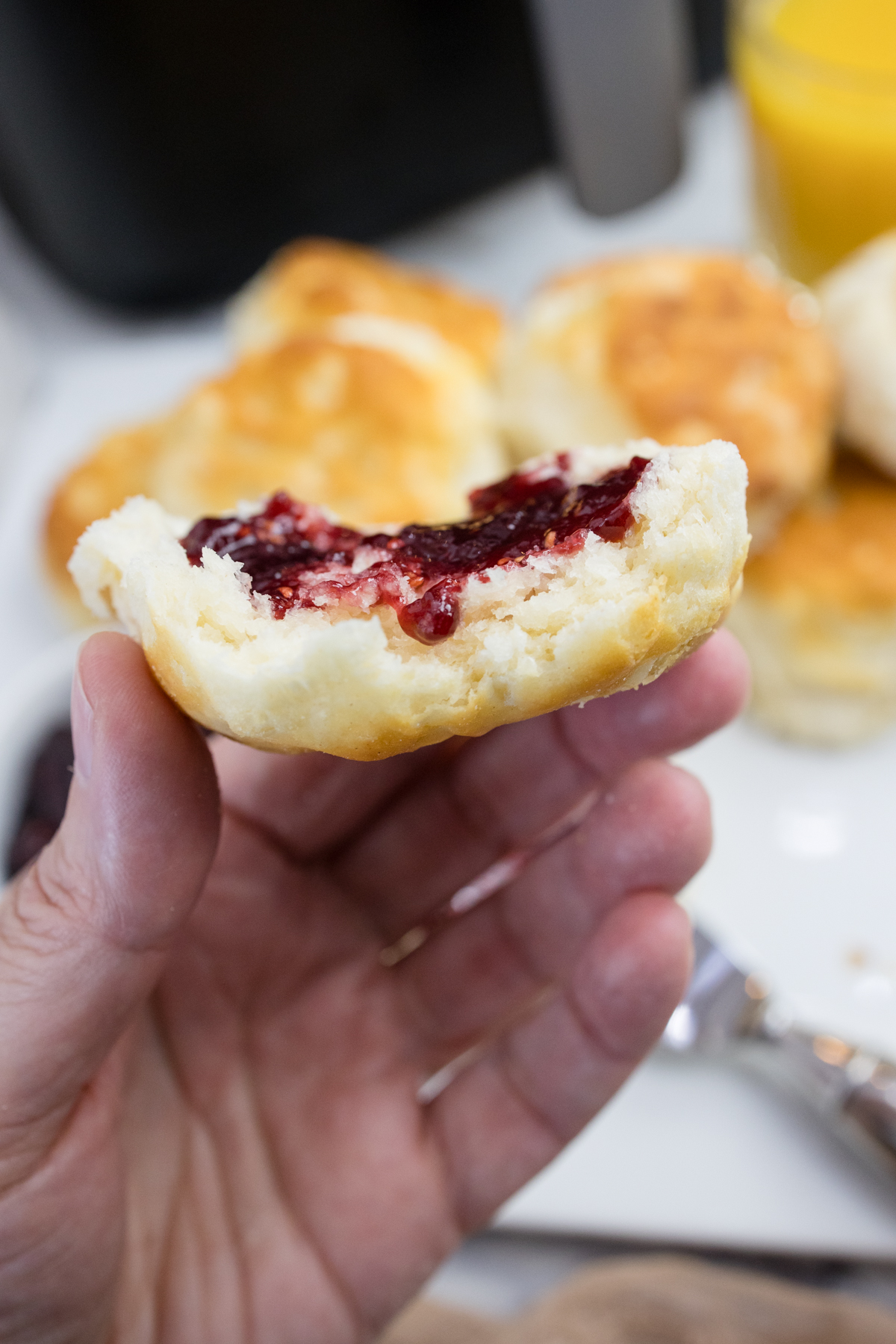 Close view of half of a biscuit with jam spread on it being held up by a hand in front of a surface with more biscuits on it.