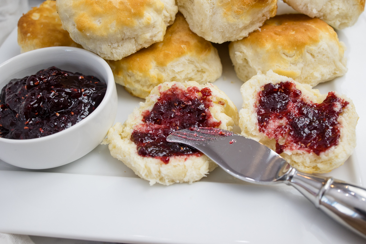 Close view of cooked biscuits on a white surface next to a ramakin of jam, and a knife is spreading jam onto a sliced biscuit.