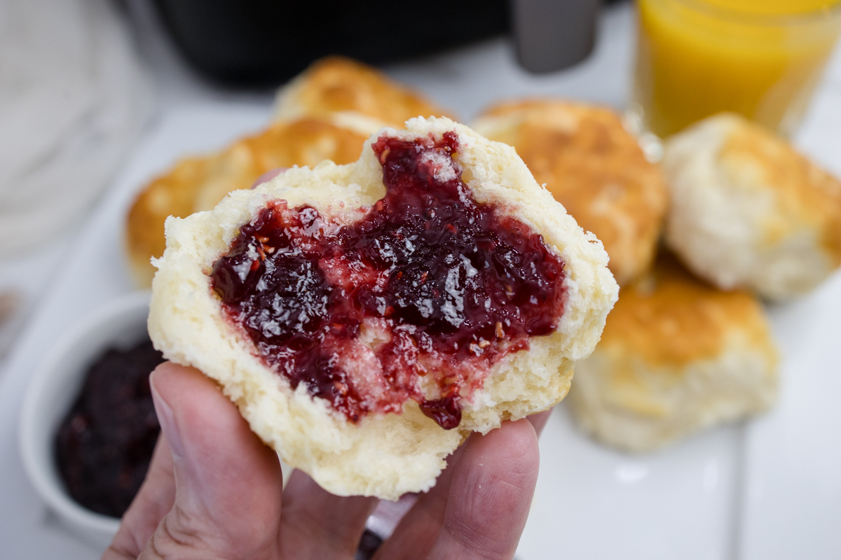 Close view of half of a biscuit with jam spread on it being held up by a hand in front of a surface with more biscuits on it.