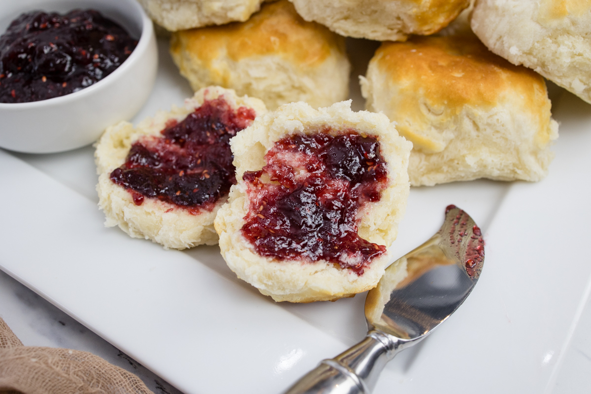 Close view of cooked biscuits on a white surface next to a ramakin of jam and a knife.