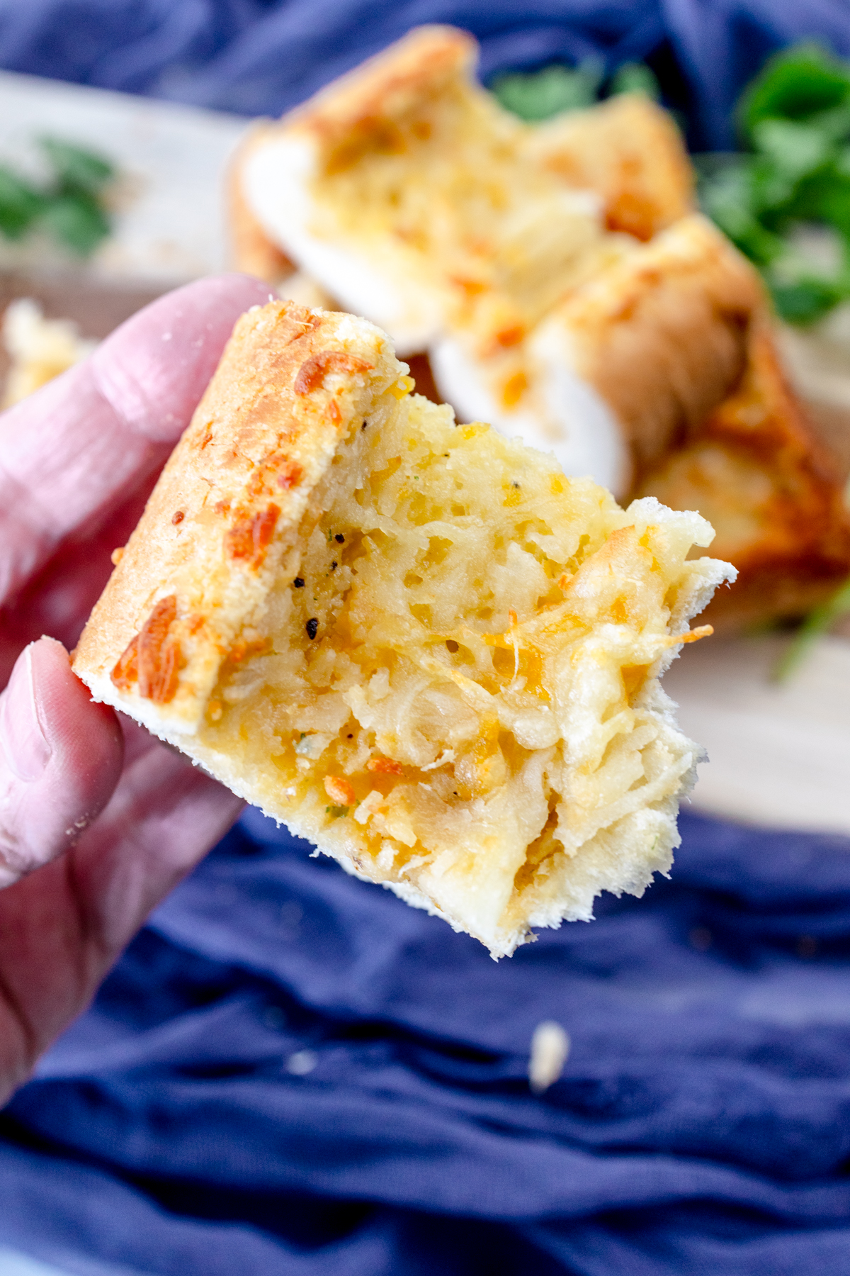 Close up image of a hand holding a slice of air fried garlic bread in front of a wooden chopping board with more garlic bread on it and fresh parsley around it for decoration.