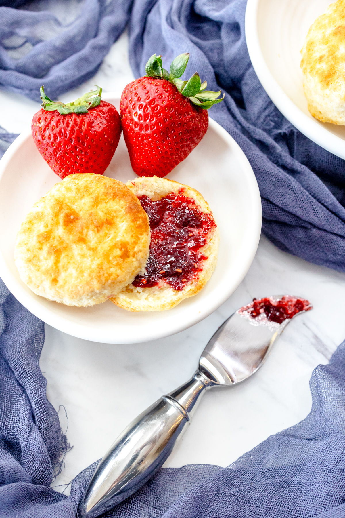 Medium-close view of a sliced cooked Air Fryer Biscuit with red jam spread on it, on a white plate with strawberries on the side, next to a knife with jam on it.