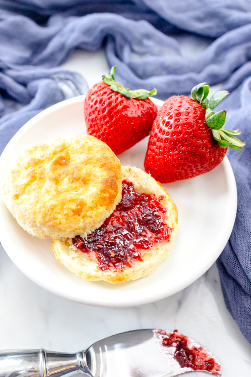 Medium-close view of a sliced cooked Air Fryer Biscuit with red jam spread on it, on a white plate with strawberries on the side, next to a knife with jam on it.