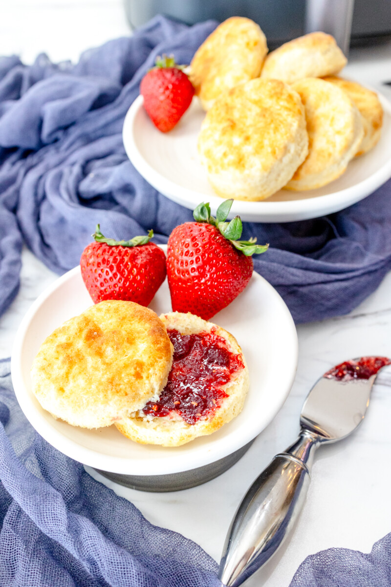 Medium-close view of a sliced cooked Air Fryer Biscuit with red jam spread on it, on a white plate with strawberries on the side, next to a knife with jam on it.