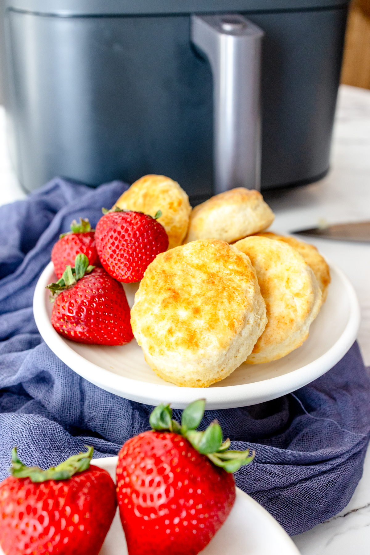 Medium-close view of cooked Air Fryer Biscuits, on a white plate with strawberries on the side, in front of an air fryer.