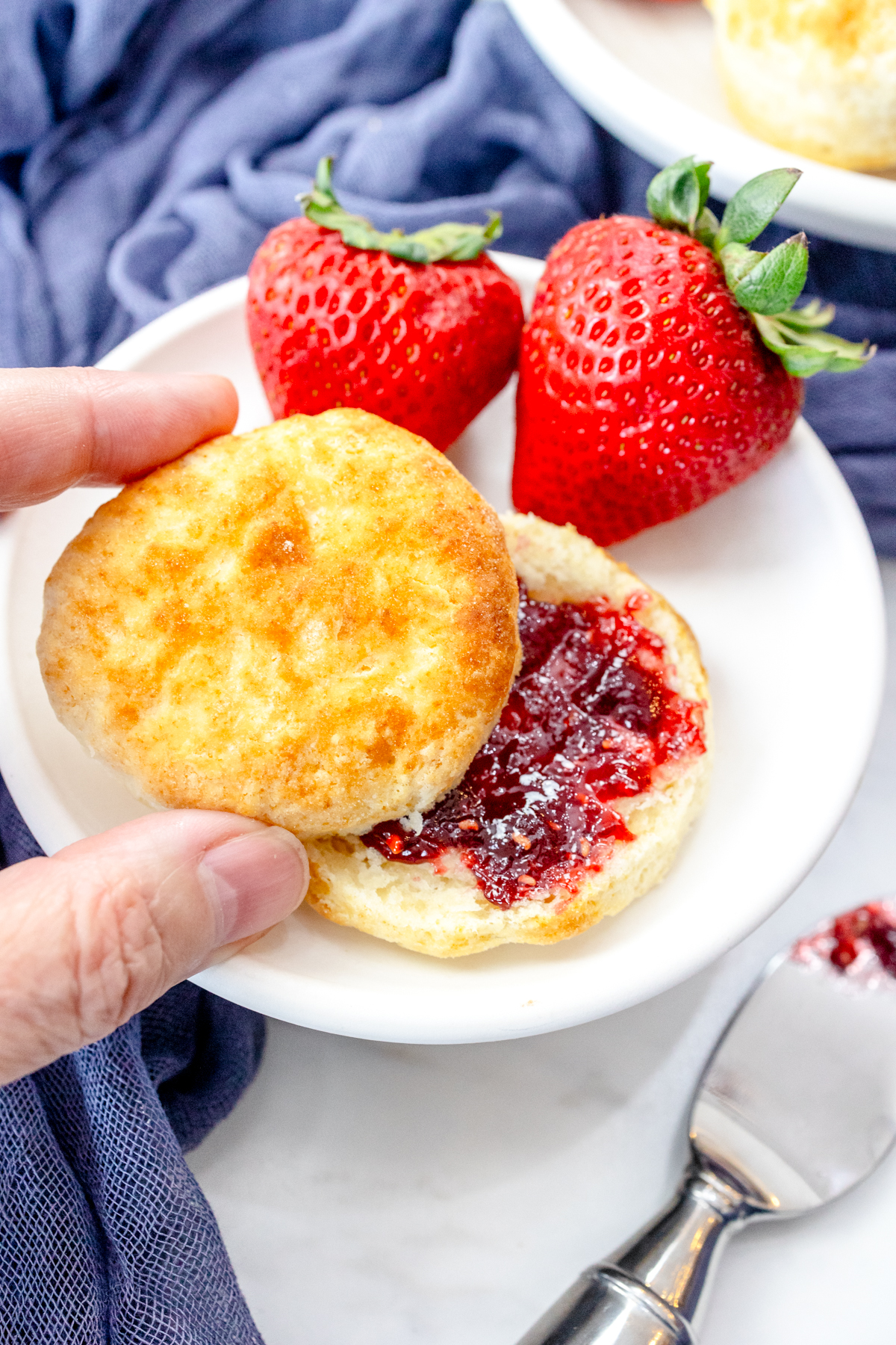 Medium-close view of a cooked Air Fryer Biscuit, on a white plate with strawberries on the side, sliced open with jam spread inside them. A hand is picking up the top of the biscuit.