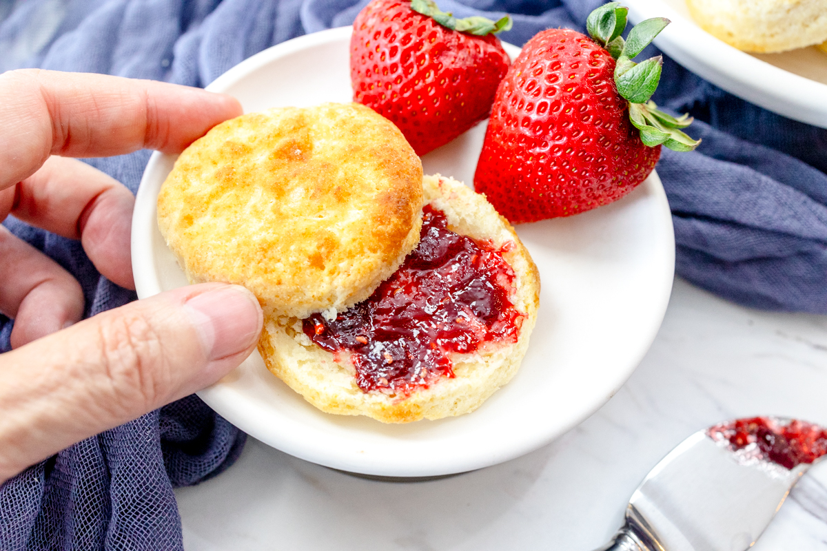 Medium-close view of a cooked Air Fryer Biscuit, on a white plate with strawberries on the side, sliced open with jam spread inside them. A hand is picking up the top of the biscuit.