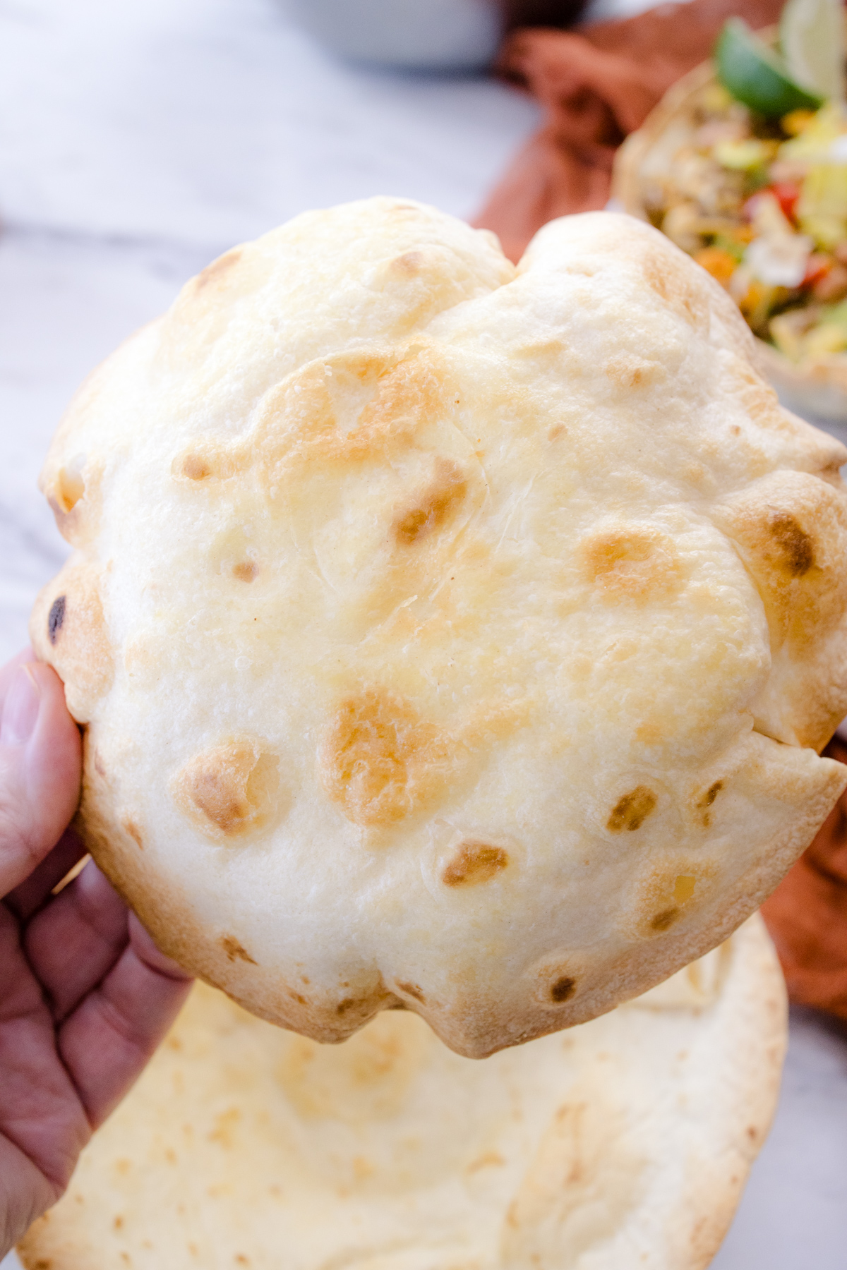 A hand holding an Air Fryer Flour Tortilla Bowl so that the bottom faces the camera, over filled tortilla bowl on a plate.