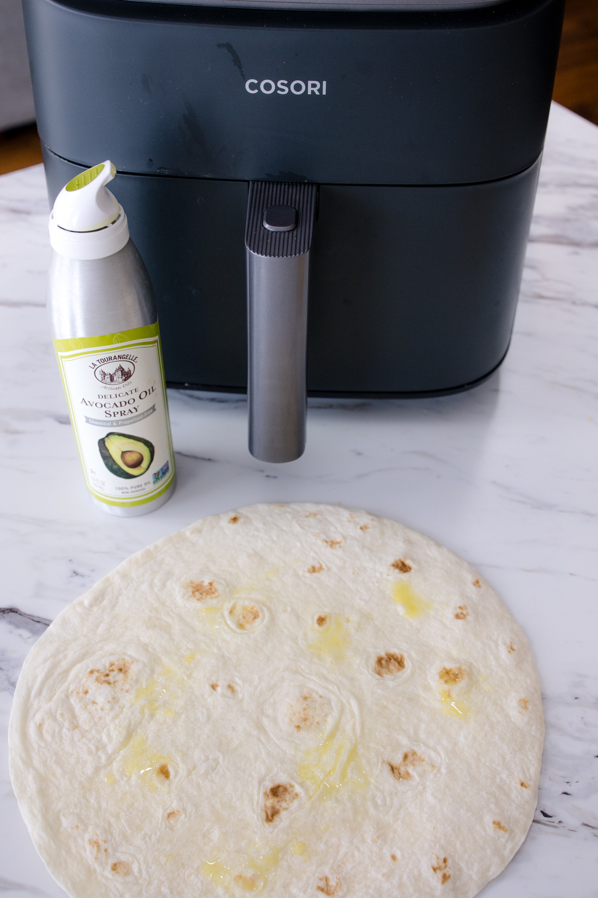 A flour tortilla sprayed with avocado oil, on a white surface, in front of an air fryer.