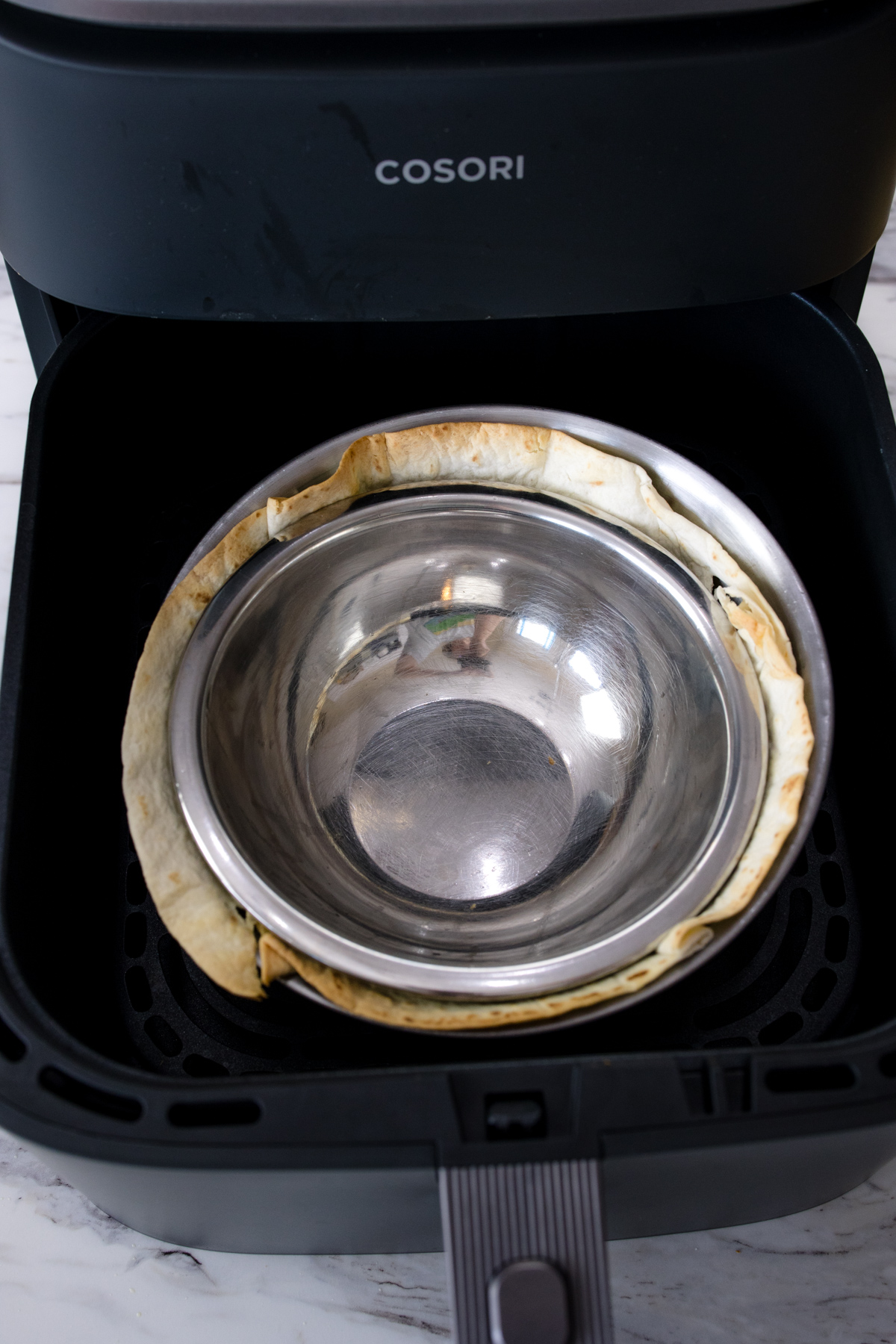 A tortilla between two metal bowls in an air fryer basket.