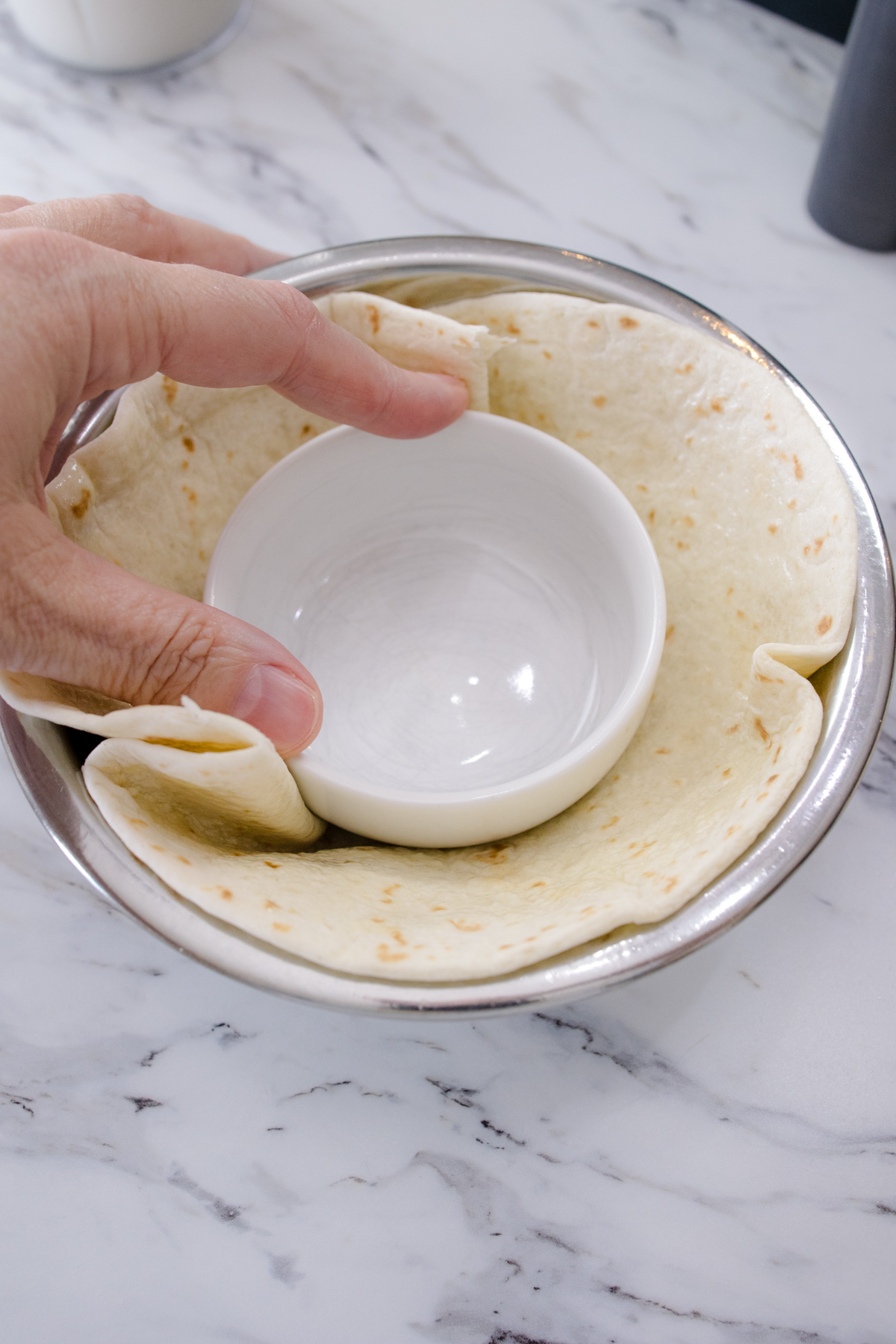 An uncooked tortilla in a metal bowl with a ramakin being pressed into the middle.