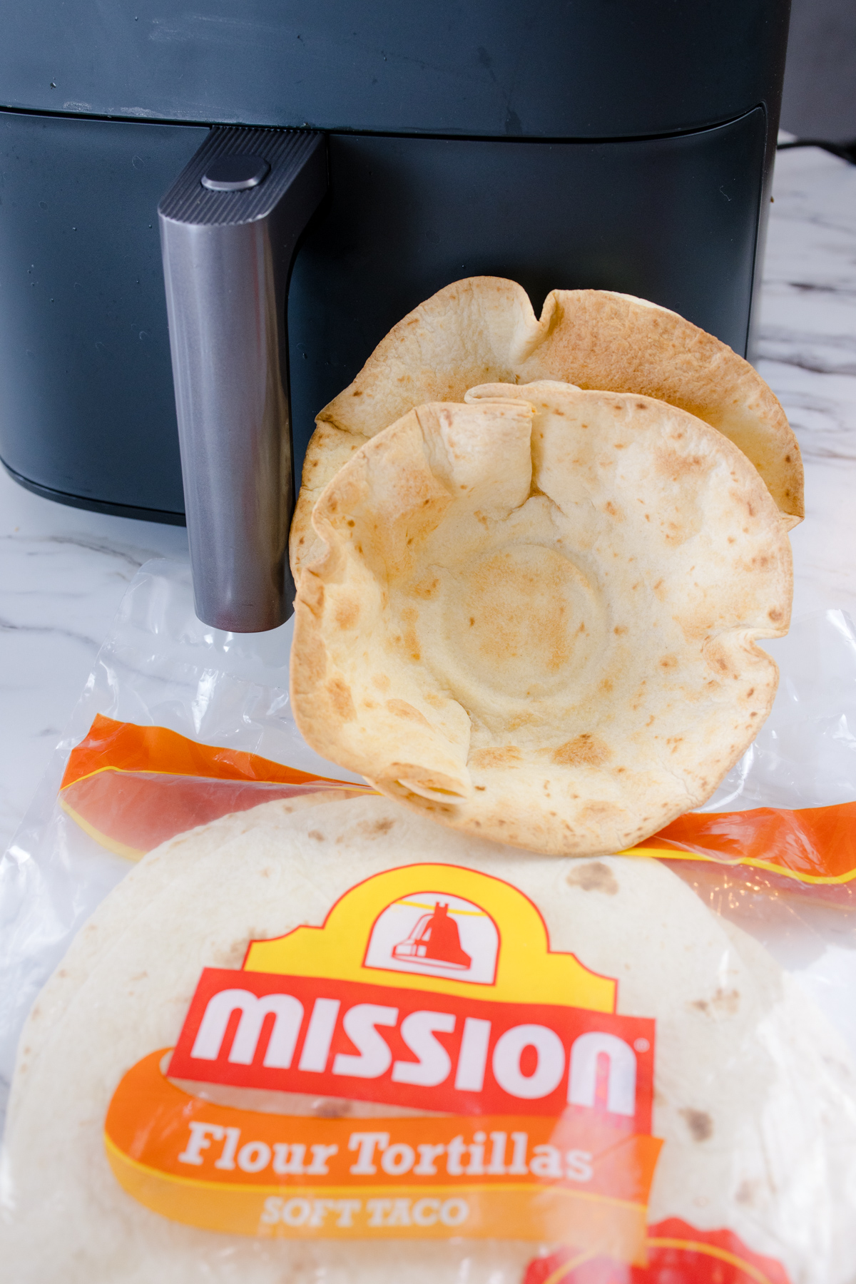 Air Fryer Flour Tortilla Bowls propped up against an air fryer with a packet of flour tortillas lying in front of them.