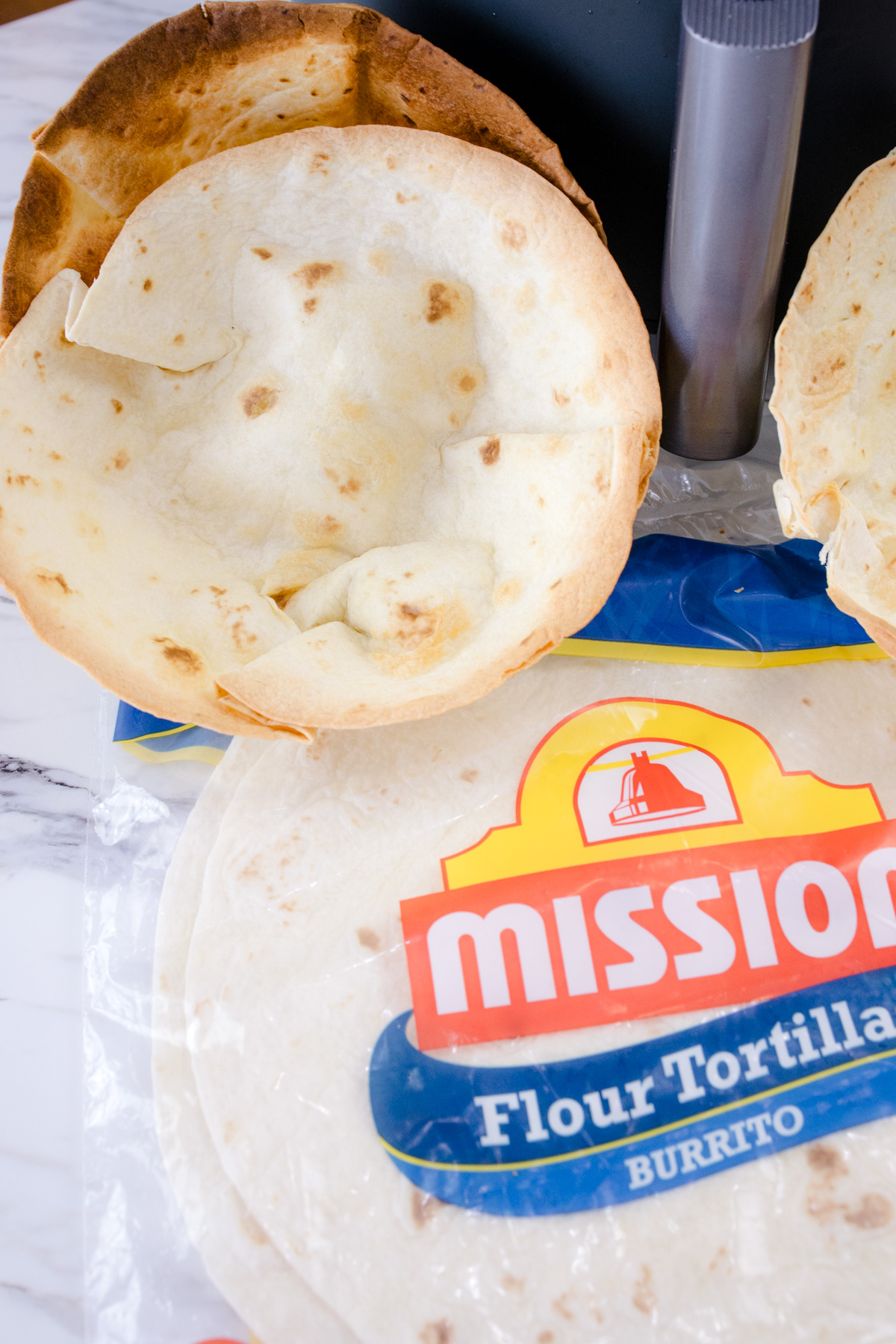 Air Fryer Flour Tortilla Bowls propped up against an air fryer with a packet of flour tortillas lying in front of them.