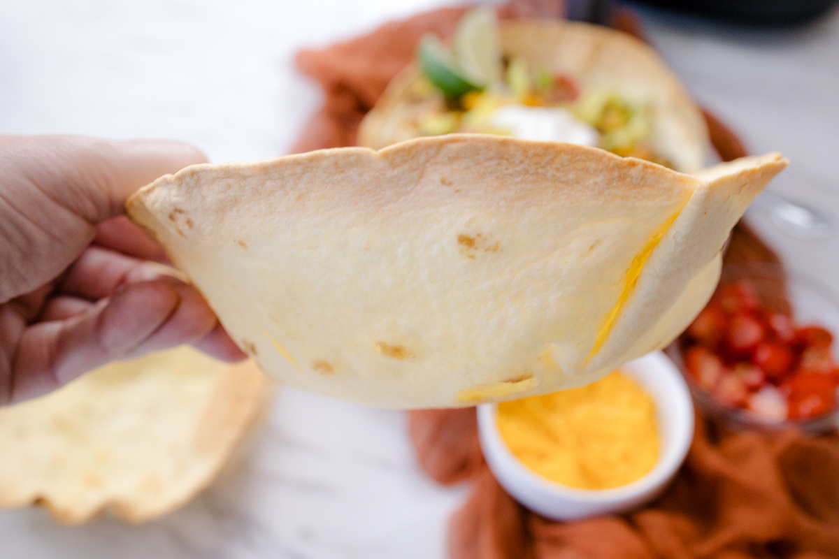 A hand holding an Air Fryer Flour Tortilla Bowl so that the side faces the camera, over filled tortilla bowl on a plate.