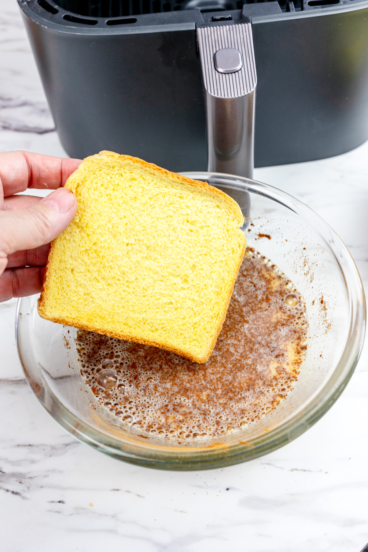 A piece of bread about to be dipped in the French Toast mixture in a large glass mixing bowl.