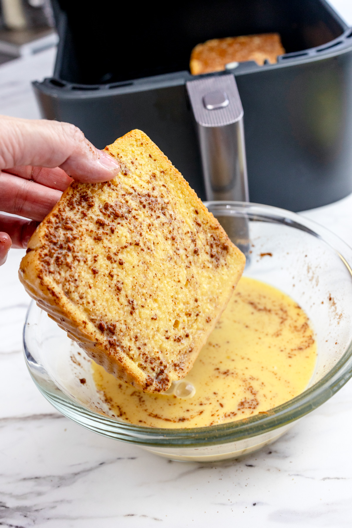 A piece of bread being dipped in the French Toast mixture in a large glass mixing bowl.