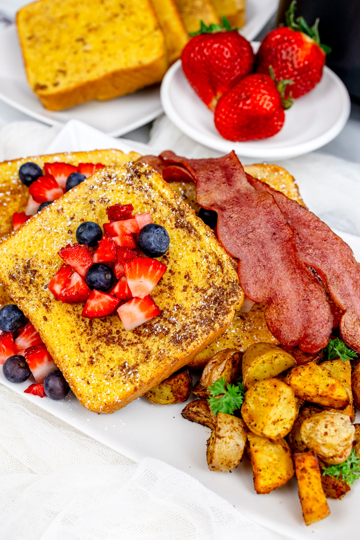 Air Fryer French Toast with syrup and berries on top on a rectangular white plate with bacon, breakfast potatoes, and more berries.