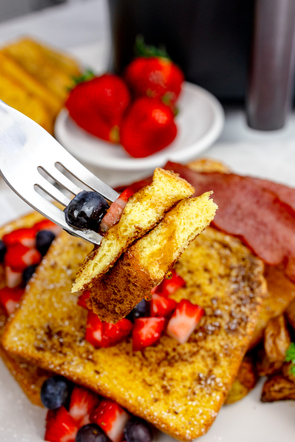 A fork picking up pieces of Air Fryer French Toast with syrup and berries on top, from a rectangular white plate with more toast and berries on it.