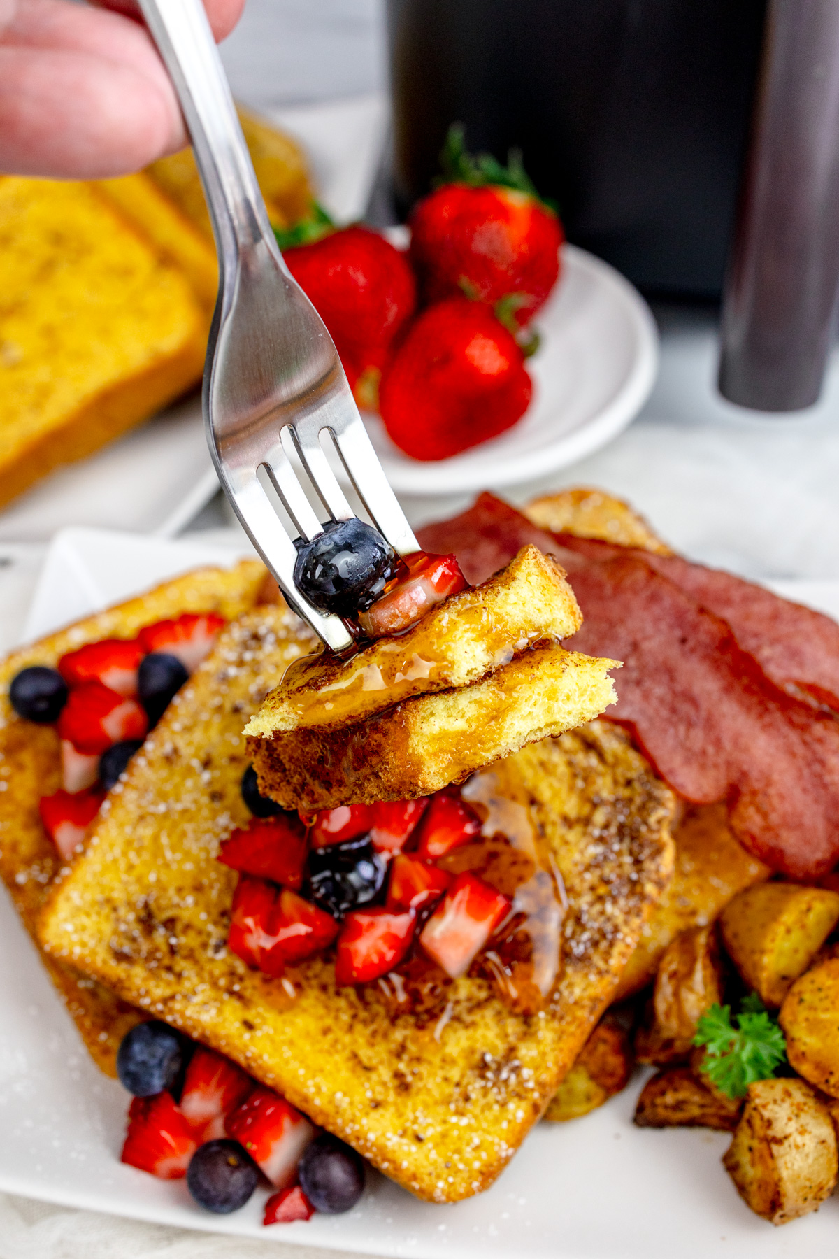 A fork picking up pieces of Air Fryer French Toast with syrup and berries on top, from a rectangular white plate with more toast and berries on it.