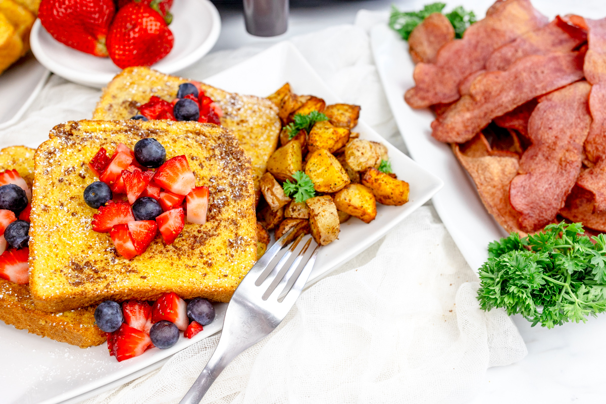 Air Fryer French Toast with syrup and berries on top on a rectangular white plate with bacon, breakfast potatoes, and more berries.