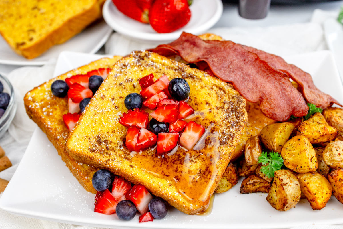 Air Fryer French Toast with syrup and berries on top on a rectangular white plate with bacon, breakfast potatoes, and more berries.