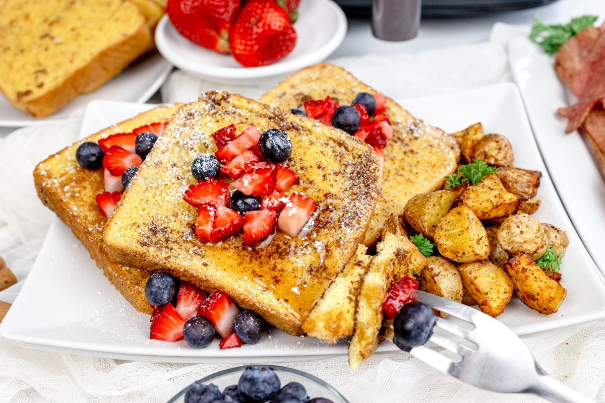 Air Fryer French Toast with syrup and berries on top on a rectangular white plate with bacon, breakfast potatoes, and more berries.