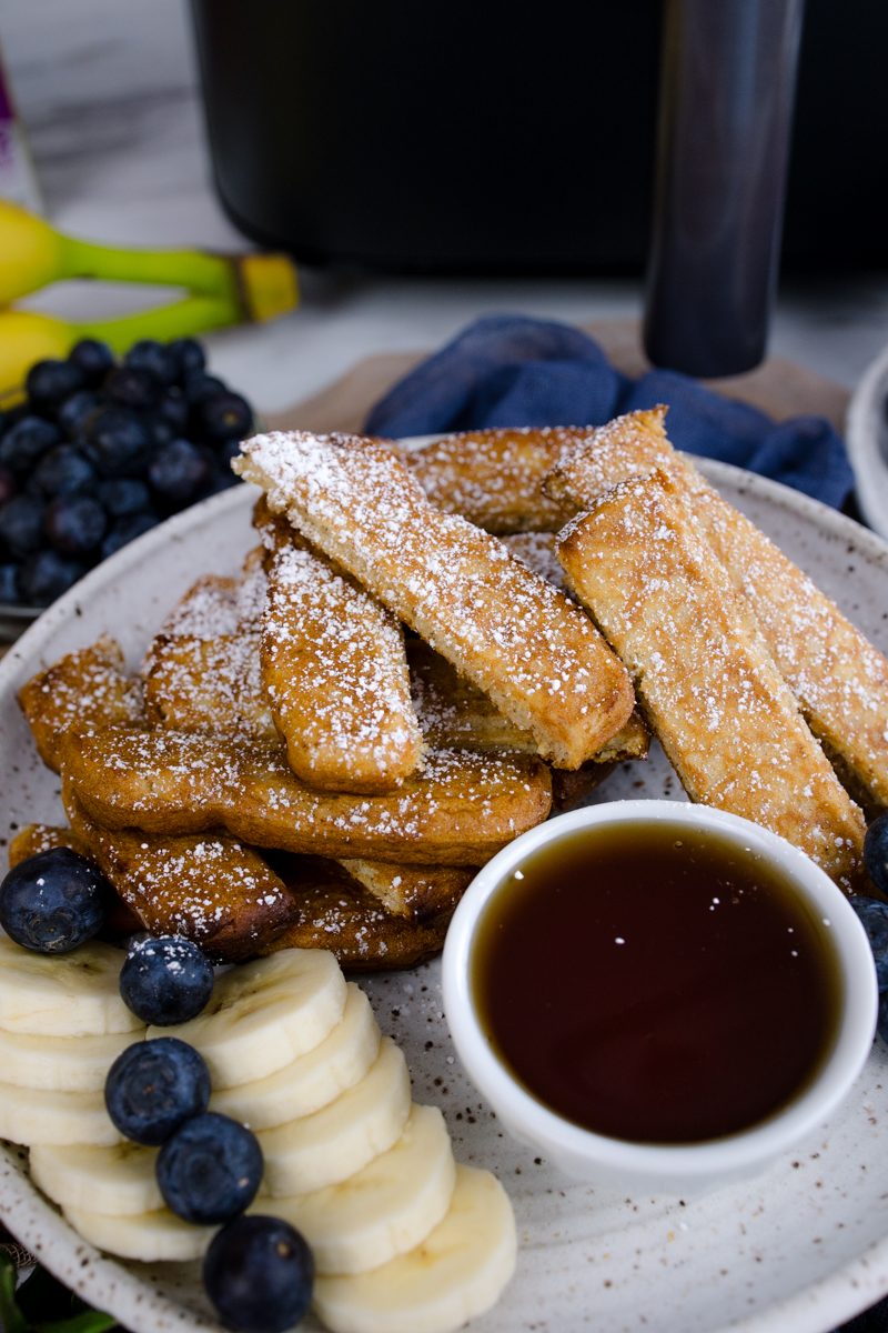 Air Fryer Frozen French Toast Sticks on a white plate with blueberries, slices of banana, and a ramekin of maple syrup.