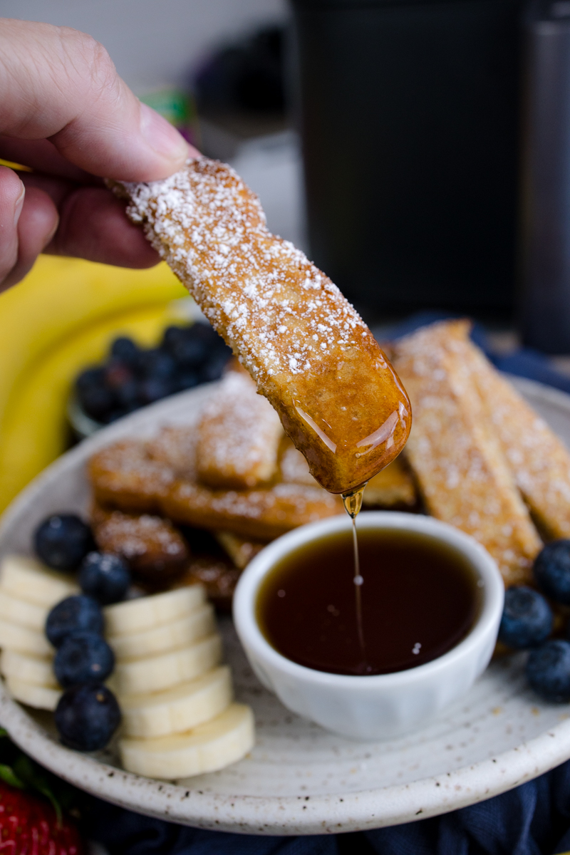 Air Fryer Frozen French Toast Sticks on a white plate with blueberries, strawberries, slices of banana, and a ramekin of maple syrup. A hand is dipping a French Toast stick into the syrup.