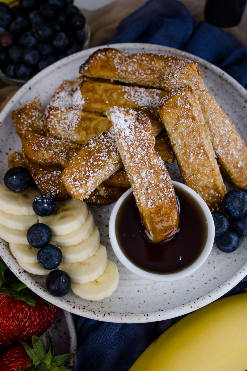 Air Fryer Frozen French Toast Sticks on a white plate with blueberries, slices of banana, and a ramekin of maple syrup.