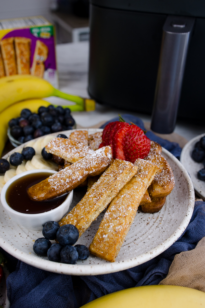 Air Fryer Frozen French Toast Sticks on a white plate with blueberries, strawberries, slices of banana, and a ramekin of maple syrup.