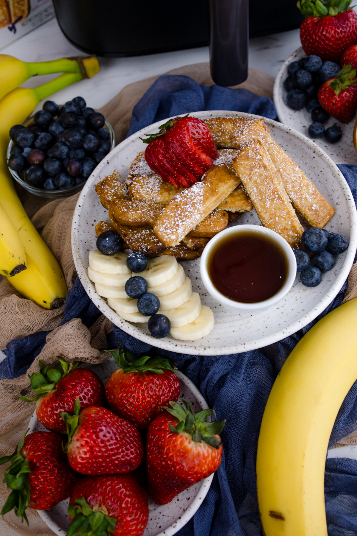 Air Fryer Frozen French Toast Sticks on a white plate with blueberries, strawberries, slices of banana, and a ramekin of maple syrup.