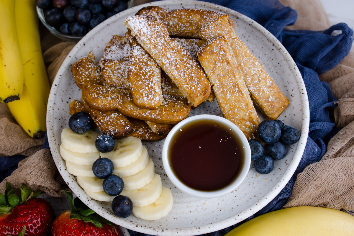 Air Fryer Frozen French Toast Sticks on a white plate with blueberries, slices of banana, and a ramekin of maple syrup.