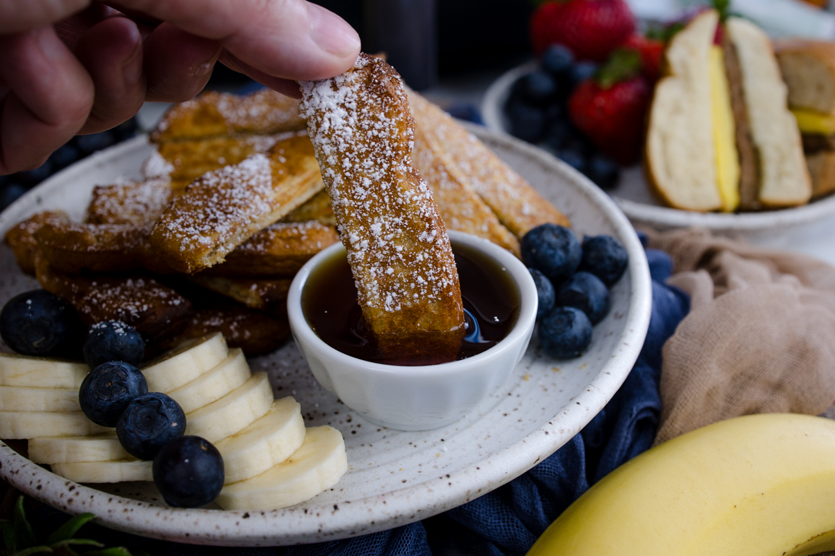 Air Fryer Frozen French Toast Sticks on a white plate with blueberries, strawberries, slices of banana, and a ramekin of maple syrup. A hand is dipping a French Toast stick into the syrup.