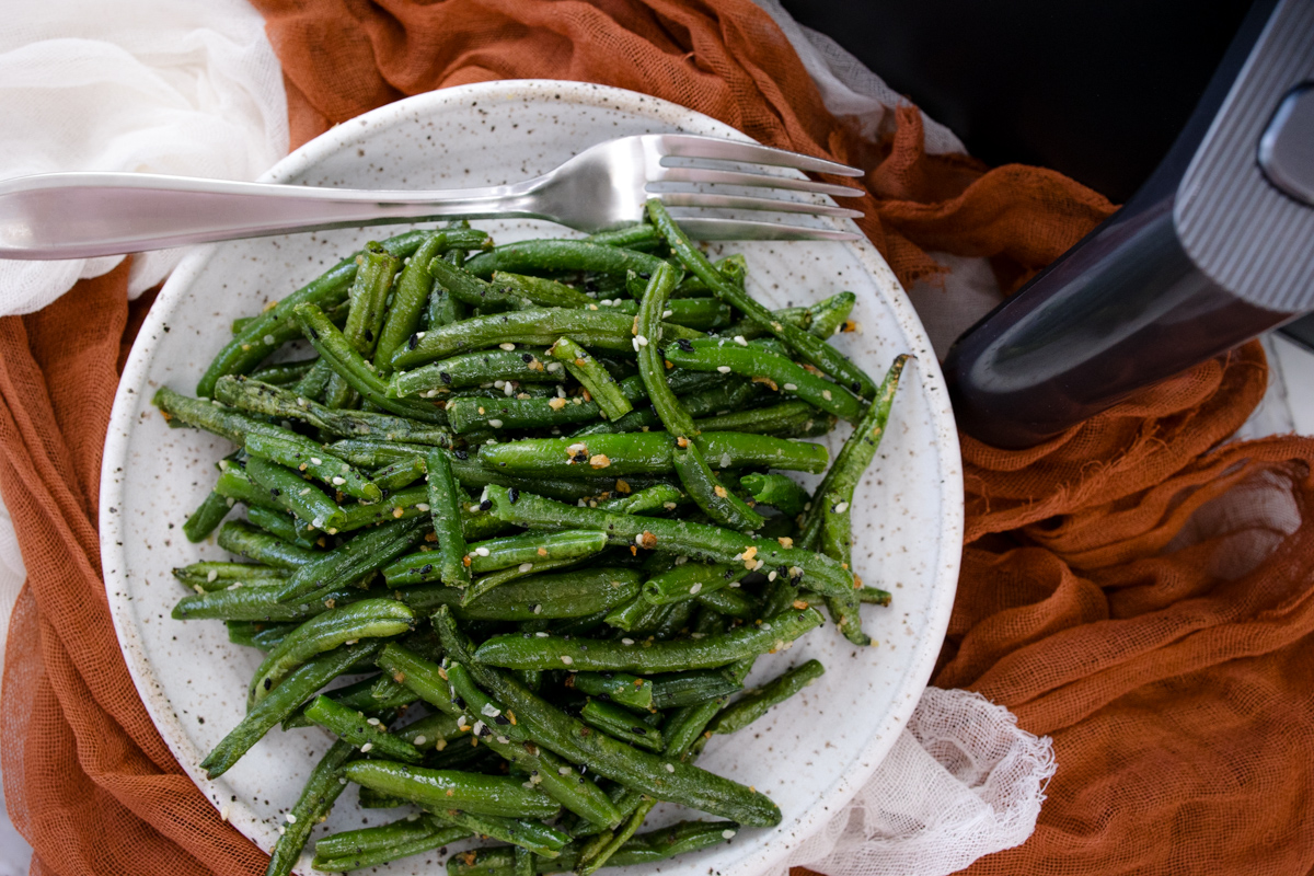 A white plate full of air fried green beans in front of an air fryer.