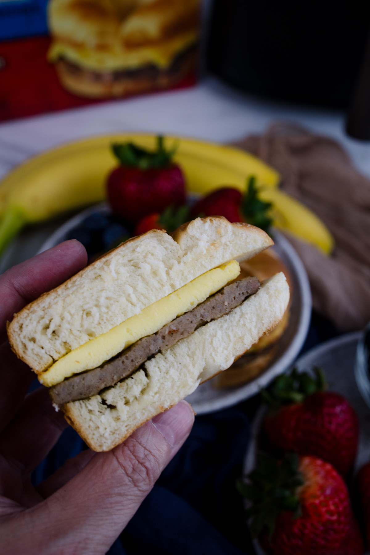 A hand holding half of a sliced Jimmy Dean Breakfast Sandwich in front of a plate with strawberries and blueberries on it.