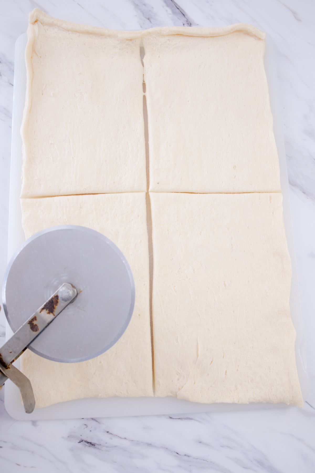 Crescent roll dough stretched in a rectangle on a white surface, being sliced with a pizza cutter.