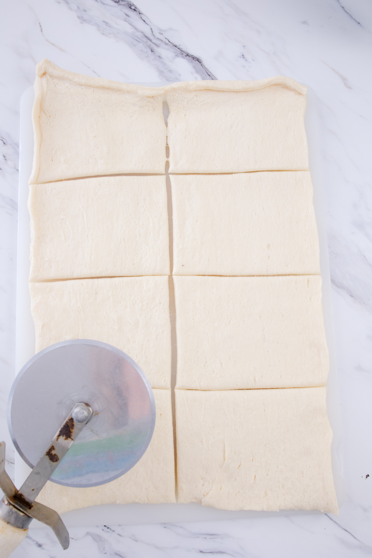 Crescent roll dough stretched in a rectangle on a white surface, being sliced with a pizza cutter.