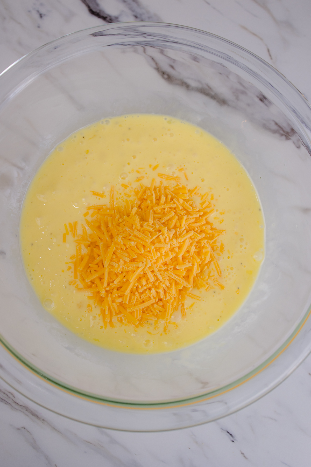 A top view of whisked eggs and milk in a glass mixing bowl with grated cheese in it.